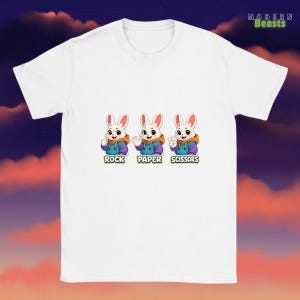 May include: White t-shirt featuring three cartoon bunnies playing rock, paper, scissors. Each bunny wears a blue hoodie with an orange hood and has a different hand gesture. The words "Rock", "Paper", and "Scissors" are below each bunny.