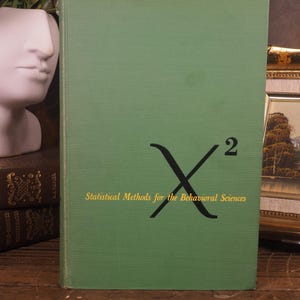 May include: A green hardcover book titled Statistical Methods for the Behavioral Sciences with a large black chi-squared symbol printed on the front cover. The text is written in a yellow serif font centered below the black symbol.