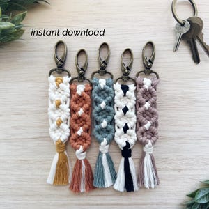 May include: Five macrame keychains in various colors, including white, rust, teal, black, and mauve, each with a coordinating tassel. The keychains have bronze-colored clasps and are displayed on a light wood surface. Text in the image reads "instant download."