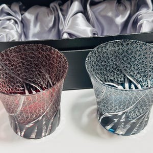 以下が含まれることがあります： 複雑な幾何学模様の装飾用グラス2個。グラス1個は赤、もう1個は青です。グラスは、銀色のサテンの裏地が付いた黒い箱の前にあります。グラスはユニークで芸術的なデザインです。