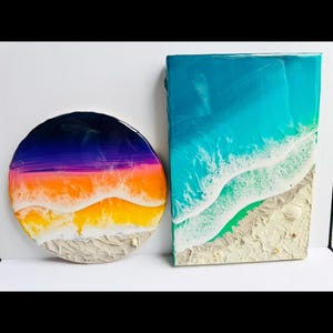 以下が含まれることがあります： ビーチの風景を描いた2つの樹脂アート作品。1つは、白い砂浜の上に紫、オレンジ、黄色の夕日のグラデーションがある丸い作品です。もう1つは、ターコイズブルーの水と白い波がある長方形の作品です。