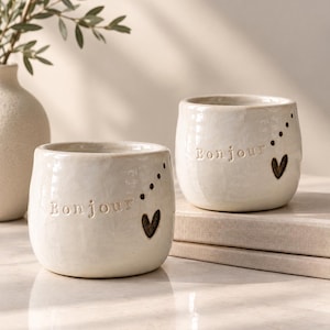 May include: Two off-white ceramic mugs with a glossy finish. Each mug features the embossed word Bonjour, a small dark heart shape, and a series of five small dots arranged in a diagonal line. The mugs have a rounded, slightly irregular shape.
