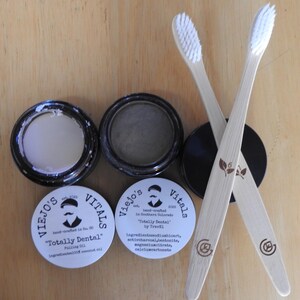May include: An overhead shot of dental care products. Two bamboo toothbrushes with white bristles are crossed. Two small jars of dental products are open, and two are closed. The jars are labeled "Viejo's Vitals" and "Totally Dental."