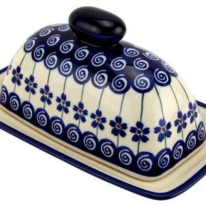 May include: A ceramic butter dish with a domed lid and a rectangular base. The dish is cream-colored with a blue floral and swirl design. The lid has a dark blue knob. The base and lid are trimmed with a dark blue border.