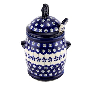 May include: A blue and white ceramic sugar bowl with a lid and a small spoon. The bowl has a floral pattern and two small handles. The lid has a decorative handle. The spoon rests on the edge of the bowl.