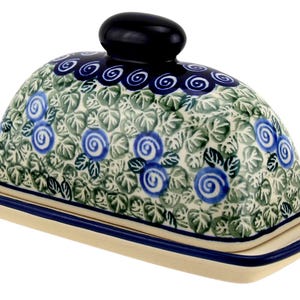 May include: Ceramic butter dish with a domed lid and rectangular base. The lid features a blue and green floral pattern with a dark blue knob. The base is cream-colored with a dark blue trim. A decorative kitchenware item.