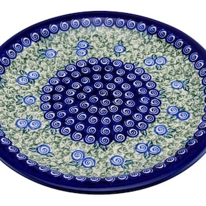 May include: A round ceramic plate featuring a central cluster of dark blue hand-painted swirls surrounded by a field of light green leaves and small blue floral accents. The rim is decorated with a continuous border of dark blue spirals.