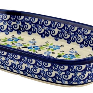 May include: A rectangular ceramic baking dish with a blue floral pattern and scrollwork. The dish has a cream-colored interior with blue flowers and green leaves. The rim and handles are dark blue.