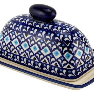 May include: A ceramic butter dish with a domed lid and rectangular base. The lid is adorned with a blue and white geometric pattern, and a small blue knob. The base is off-white with a blue trim.