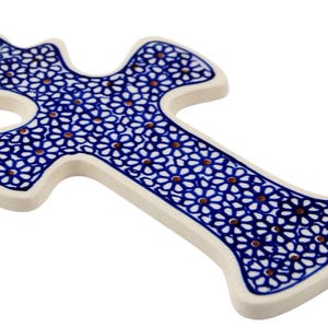 May include: A ceramic cross with a blue floral pattern on a white background. The cross has a hole at the top for hanging. The design features small white flowers with brown centers.