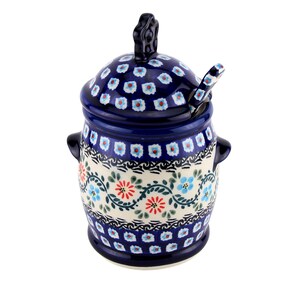 May include: A blue and white ceramic sugar bowl with a lid and a small spoon. The bowl has a floral pattern with red, blue, and green accents. The lid has a handle and a matching floral design. The spoon rests on the lid.