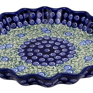 May include: A round, ceramic pie dish with a wavy edge. The dish features a dark blue rim and a decorative pattern of blue flowers and white swirls on a green background. The center of the dish has a dark blue spiral design.