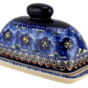 May include: A ceramic butter dish with a domed lid and a rectangular base. The dish is decorated with blue floral patterns and a dark blue knob on top. The base has a cream color.