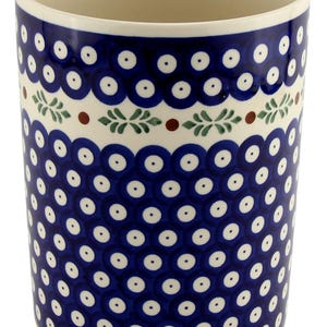 May include: A cylindrical ceramic mug with a blue and white pattern. The design features a repeating pattern of blue circles with white dots, and a band of green leaves and red dots. The interior is a cream color.