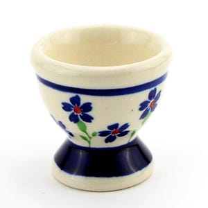 May include: A ceramic egg cup with a cream-colored base, adorned with blue floral patterns and a dark blue band. The base features a dark blue pedestal design. The egg cup is designed to hold a single egg.