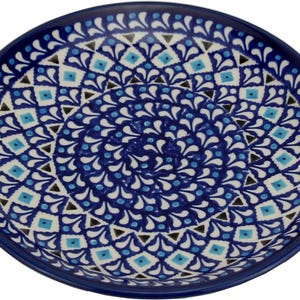 May include: A round ceramic plate with a deep blue rim and a detailed, symmetrical pattern. The design features a central spiral of white, light blue, and dark blue shapes, creating a decorative and artistic look. The plate is suitable for serving food.