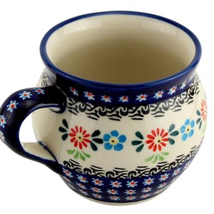 May include: A ceramic mug with a rounded shape and a handle. The mug features a cream-colored base adorned with a floral pattern in red, blue, and green. The rim and handle are detailed with a dark blue trim.