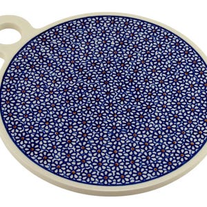 May include: Round ceramic serving board with a handle. The board features a blue and white floral pattern with a cream-colored rim and handle. The floral design is a repeating pattern of small, stylized flowers.
