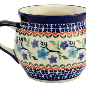 May include: A ceramic mug with a rounded shape and a dark blue handle. The mug features a floral pattern with blue flowers, green leaves, and orange accents on a cream background. The rim is dark blue.