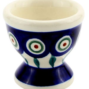 May include: A ceramic egg cup with a white interior and rim. The exterior features a dark blue band with a repeating pattern of green and white circles with red centers. The base is also dark blue.