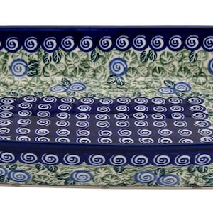 May include: Rectangular ceramic baking dish with a dark blue rim and floral pattern. The dish features a repeating pattern of blue flowers and green leaves on a white background. The edges are decorated with a swirl design.