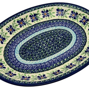 May include: An oval ceramic platter featuring a cream-colored base with a decorative border of blue flowers and green leaves. The center of the platter displays a dense, intricate pattern of small blue and green floral motifs surrounded by a blue rim.