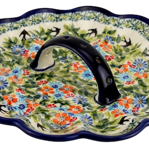 May include: Ceramic serving dish with a scalloped edge and a dark blue handle. The dish features a floral pattern with blue, orange, and green flowers, and black bird silhouettes. The rim is dark blue.