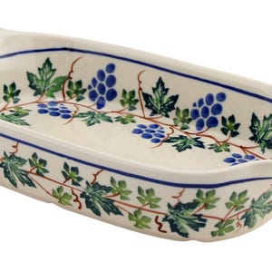 May include: Cream-colored ceramic serving dish with a grape and leaf design. The oval dish has a blue border and handles. The grape clusters are blue, and the leaves are green.