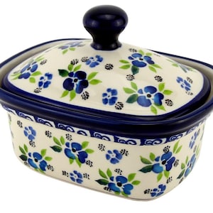 May include: A ceramic butter dish with a lid. The dish and lid are cream-colored and decorated with blue floral patterns and green leaves. The rim and the knob on the lid are dark blue.