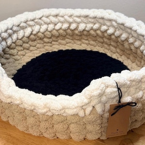 May include: A round, hand-crocheted pet bed made of thick, chunky yarn. The exterior features a two-tone design with a beige base and a cream-colored rim. The interior floor of the bed is solid black. A brown paper tag is attached to the rim.