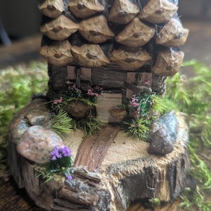 May include: A miniature house sculpture crafted from natural wood and pinecone scales. The roof features layered pinecone scales, while the base is a circular wood slice decorated with smooth river stones, small green moss, and tiny purple flowers.