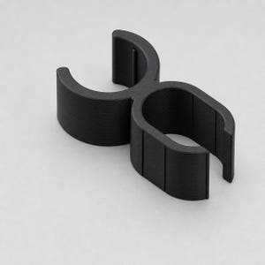 May include: A matte black plastic double clip connector featuring two C-shaped openings designed to hold two cylindrical objects side by side. The component has a textured surface and a rigid structure with vertical reinforcement ribs along the exterior.