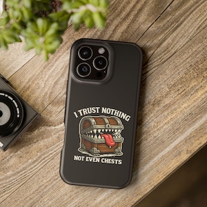 May include: Black phone case with the text "I TRUST NOTHING NOT EVEN CHESTS" and a cartoon treasure chest design with teeth and a tongue. The case is on a wooden surface.