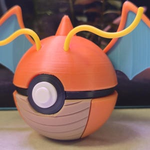 May include: A 3D-printed orange and tan ball-shaped figure with blue wings and yellow antennae. The figure has a white button and black circle on the front. The figure is a stylized representation of a character from a popular animated series.