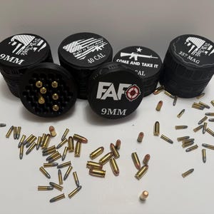 May include: Black cylindrical bullet holders with white text and graphics, including "9MM", "40 CAL", and "357 MAG". The holders are stacked and surrounded by loose bullets on a white surface. One holder is open, displaying bullet chambers.