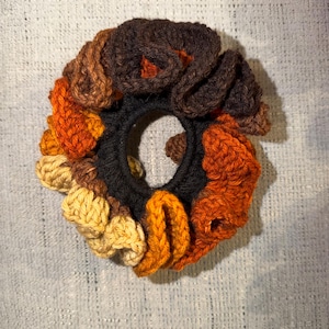 Peut inclure: Un chouchou fait à la main avec un assortiment de couleurs chaudes. La base élastique noire est ornée de fils crochetés bruns, oranges et jaunes. Cet accessoire est conçu pour attacher les cheveux.