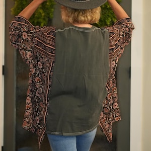May include: A gray fedora hat with a decorative band is held up by a person. The person is wearing a dark green top with a floral patterned kimono-style jacket and light blue jeans.