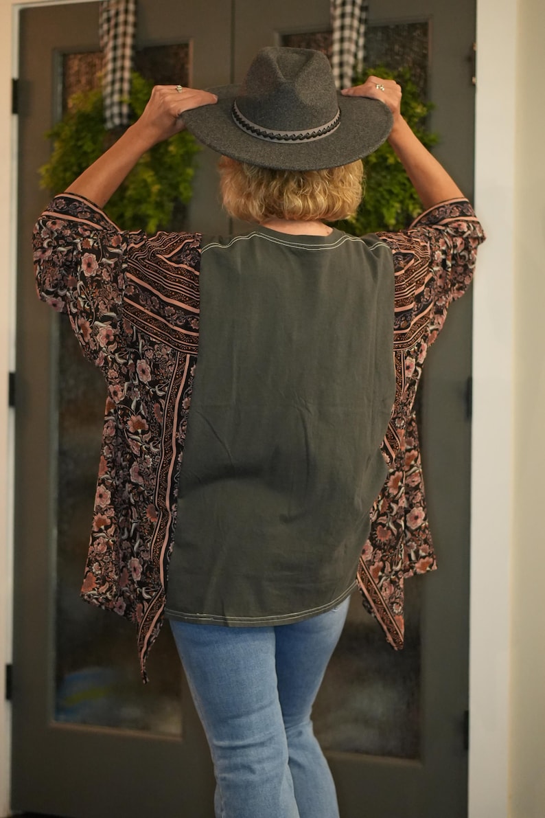 May include: A gray fedora hat with a decorative band is held up by a person. The person is wearing a dark green top with a floral patterned kimono-style jacket and light blue jeans.