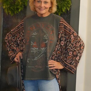 May include: A woman wearing a dark green t-shirt with a desert scene graphic and floral print sleeves. The shirt has the text "NAVY" and "MORNING". She is also wearing blue jeans and holding a gray hat.