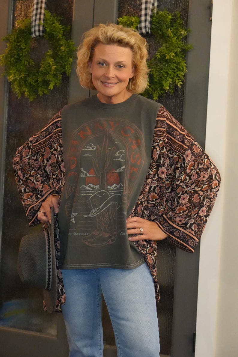 May include: A woman wearing a dark green t-shirt with a desert scene graphic and floral print sleeves. The shirt has the text "NAVY" and "MORNING". She is also wearing blue jeans and holding a gray hat.
