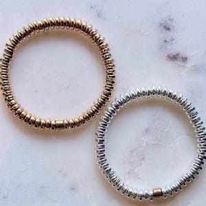 May include: Two circular stretch bracelets composed of small, metallic, disc-shaped beads. One bracelet features a gold-toned finish, while the other is silver-toned. Each bracelet includes a single, slightly larger, smooth cylindrical bead.