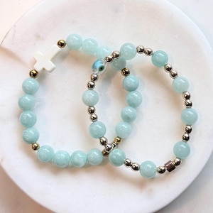 May include: Two stretch bracelets featuring light blue round beads interspersed with metallic gold-tone spheres. One bracelet includes a white cross charm, while the other features a small blue and white evil eye bead and a single dark metallic cube.