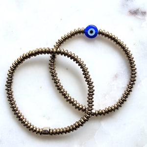 May include: Two stretch bracelets composed of small, metallic, disc-shaped beads in a bronze tone. One bracelet features a single, round, blue evil eye bead with a white and black center. The other bracelet includes a small, smooth, silver-toned bead.