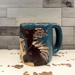 May include: A teal ceramic mug featuring a relief design of two horses with dark brown coats and light-colored manes. The mug has a large, sturdy handle and a smooth, glossy finish, showcasing detailed carvings of the horse heads and flowing manes.