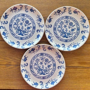 May include: Three white ceramic plates with a blue floral design. Each plate features a central floral motif surrounded by a border of stylized flowers and leaves. The plates are arranged on a wooden surface.