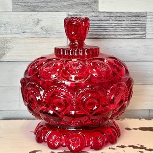May include: A ruby red glass candy dish with a lid. The dish has a round body with a starburst pattern and a pedestal base. The lid has a decorative finial. The dish is approximately 7 inches tall.