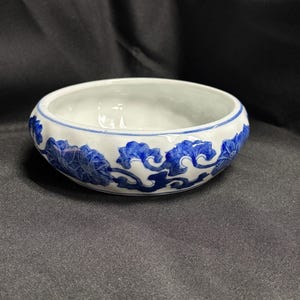 May include: A small, round, white ceramic bowl with a blue floral design around the rim. The bowl's interior is plain white, and the exterior features a continuous pattern of blue flowers and swirling leaves. The bowl is likely for decorative or serving purposes.