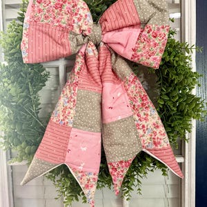 May include: A large, decorative bow made of patchwork fabric, featuring floral, striped, and polka dot patterns in shades of pink, beige, and gray. The bow is attached to a green wreath, creating a charming, handcrafted look for home decor.