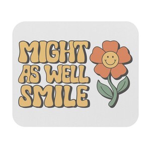 May include: A white, rectangular mousepad with the text "MIGHT AS WELL SMILE" in orange letters. A cartoon flower with an orange petal and a smiling face is on the right side of the mousepad.