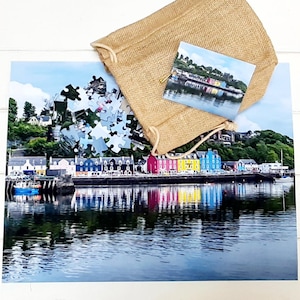Può includere: Un puzzle colorato che rappresenta una città sul mare con case dai colori vivaci e una barca blu nel porto. I pezzi del puzzle sono sparsi intorno a una borsa di iuta.